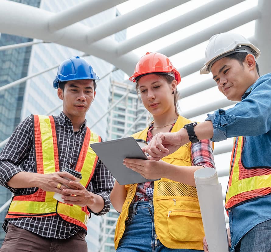 Two asian males and a woman manager looking at a tablet wearing hard hats and safety vest.