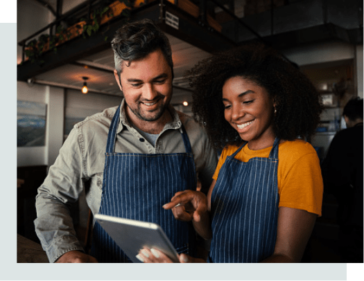 A Black female wearing a blue apron showing her white male co-worker something on her tablet. They are in a cafe setting.