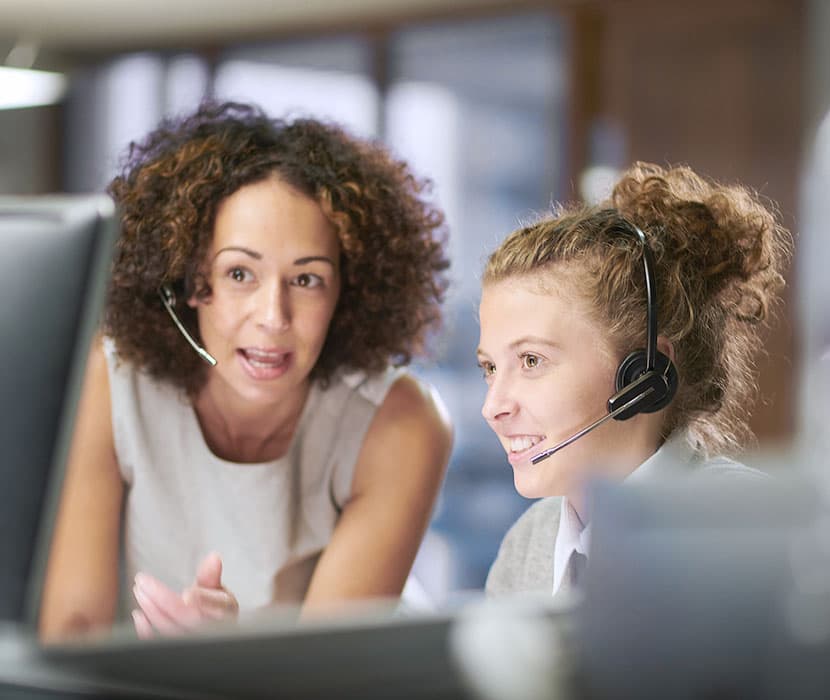 “Two female co-workers with headsets on are looking at the computer screen together.