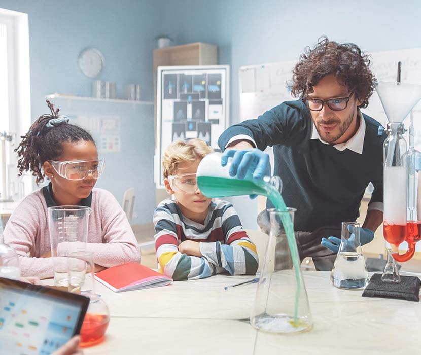 A male science teacher pouring a blue liquid in a beaker in front of elementary students in a classroom